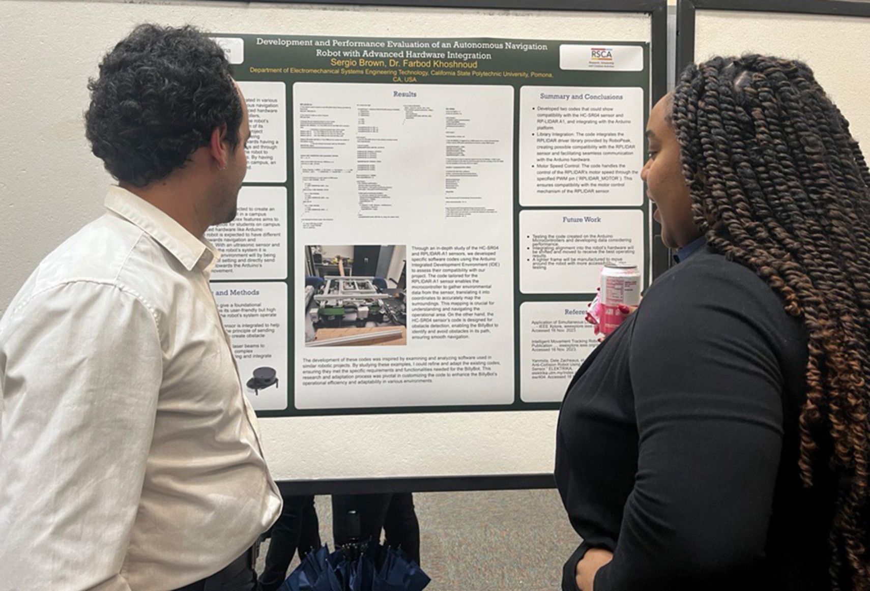 Two people are examining a research poster on autonomous navigation robots by Sergio Brown at Cal Poly Pomona. The left-side person in a white shirt is partially visible, while the right-side person with braided hair faces the poster, which includes text and images detailing the project.