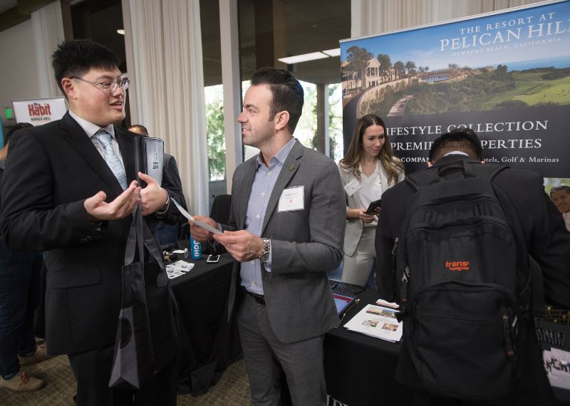 A student in business attire speaks with an employer at a career fair booth.