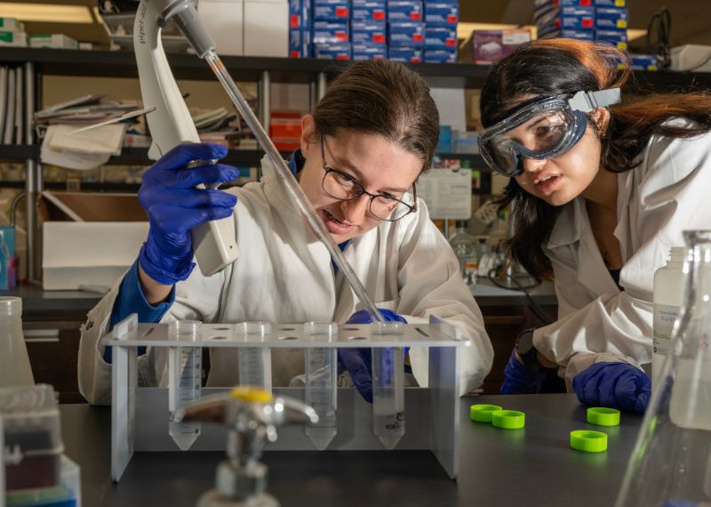 Two students in lab coats and goggles conduct an experiment using pipettes in a science laboratory.