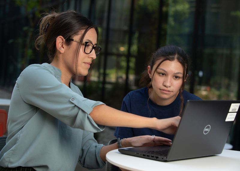 A faculty member mentors a student outdoors, guiding her through coursework on a laptop.