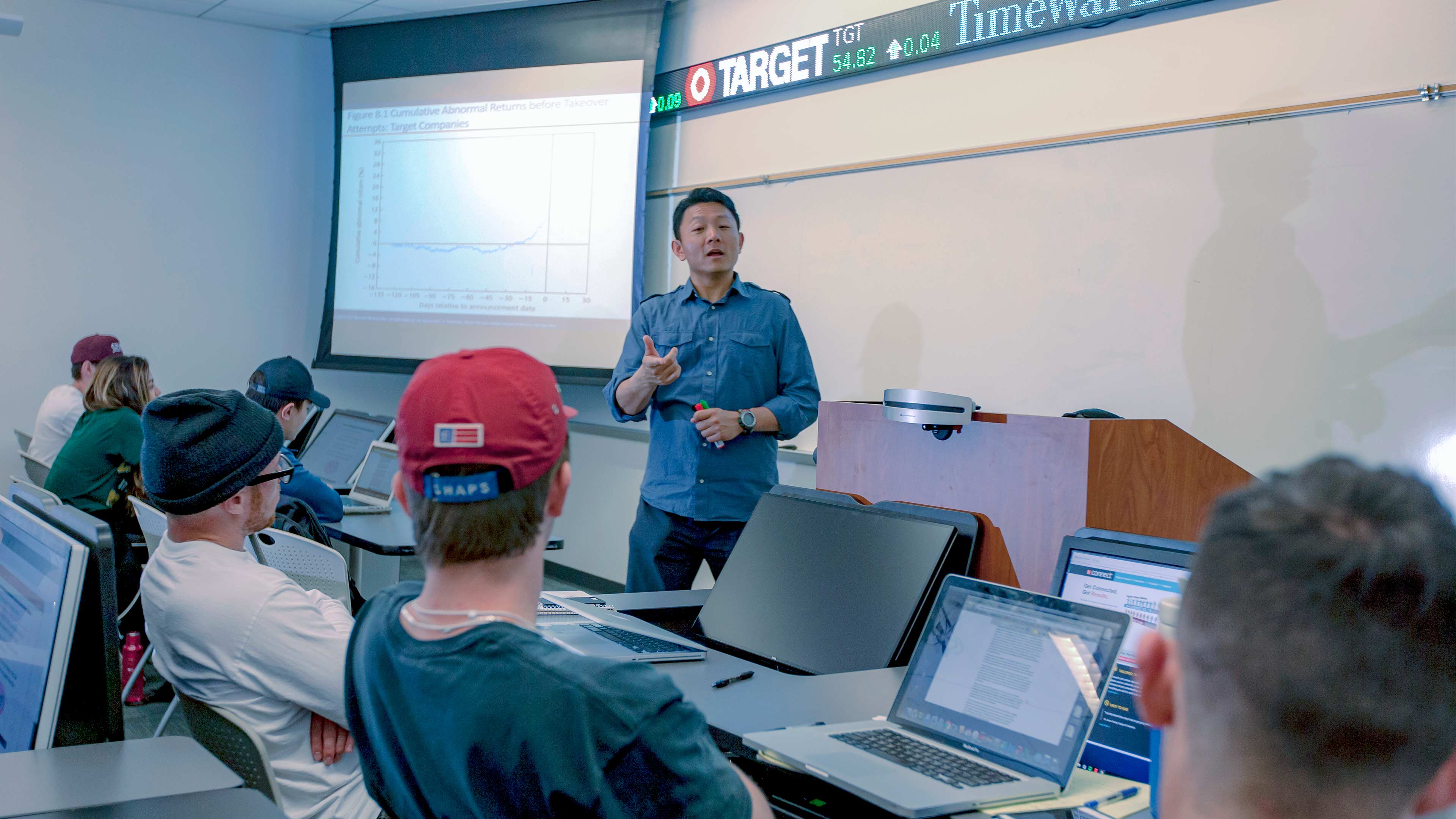 Professor in Bloomberg Terminal lab with stock ticker and lesson in background