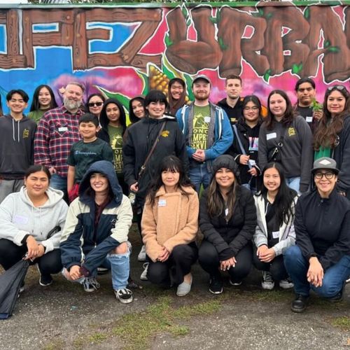 "A diverse group of individuals poses for a group photo in front of a mural that reads 'Urban Farm' during an outdoor event."
