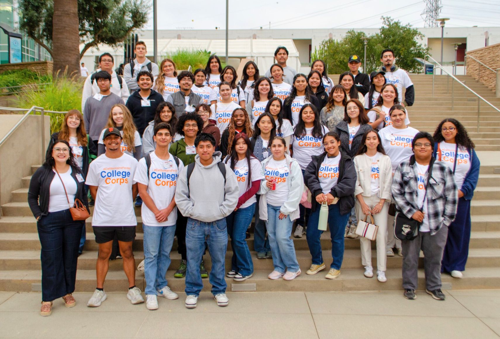 A large group of diverse College Corps members posing together in a conference room, wearing matching white t-shirts with the College Corps logo, smiling in front of a branded backdrop.