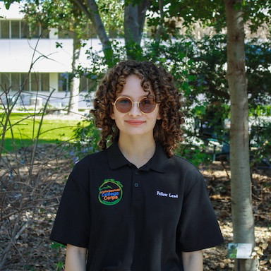 a woman with curly hair and glasses smiling in front of plants and a building