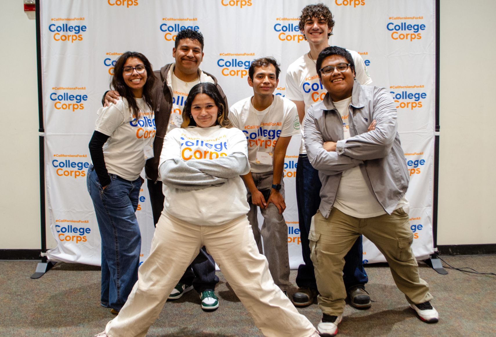 Six College Corps members posing energetically in front of a College Corps backdrop, with one person in the front striking a dynamic stance and others smiling confidently.
