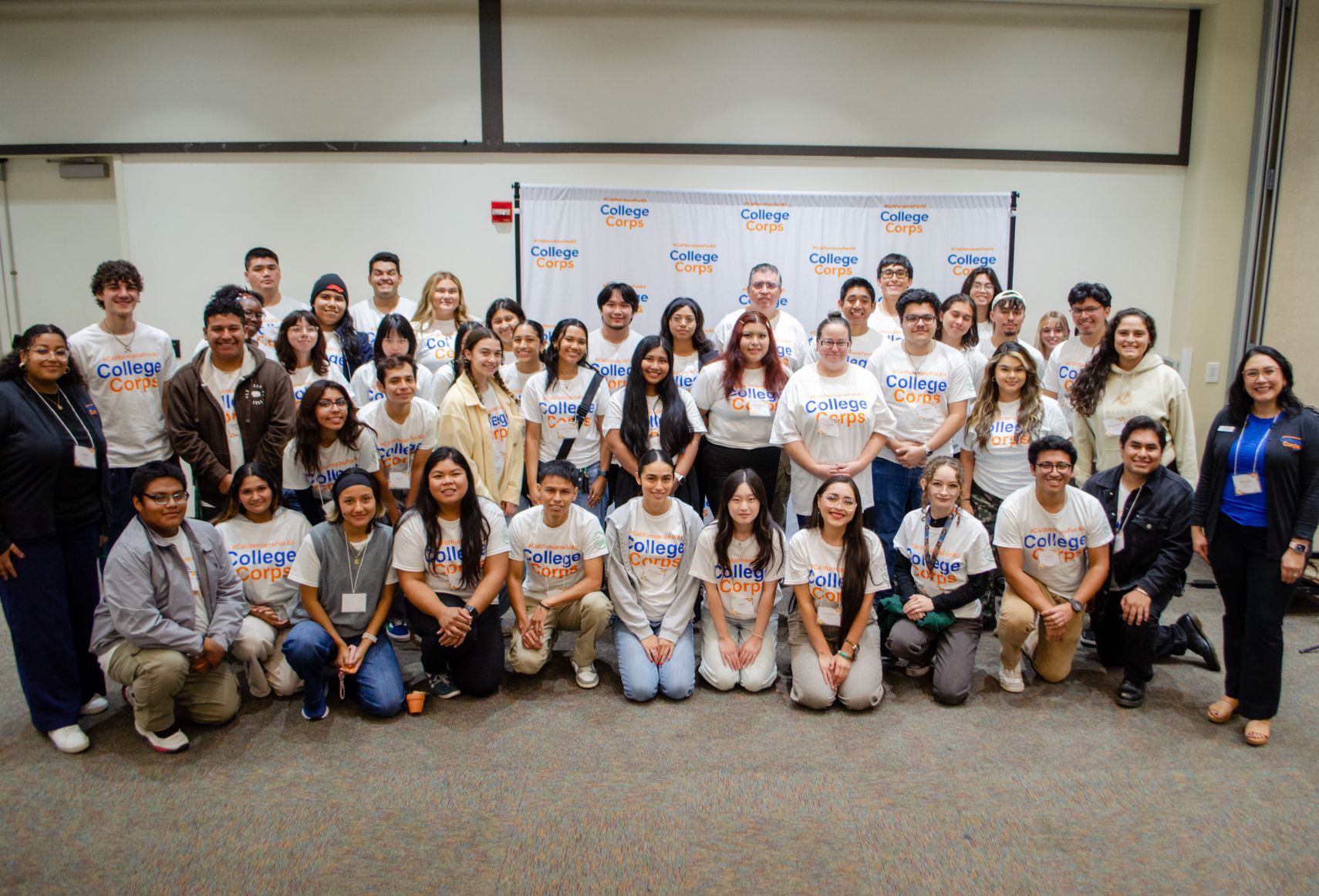 A large group of diverse College Corps members posing together in a conference room, wearing matching white t-shirts with the College Corps logo, smiling in front of a branded backdrop.