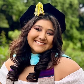Portrait of female smiling. She is wearing her tam. She has long brown hair that is curled. Nature background. 
