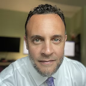 Selfie of male. He is wearing a white button up with a purple tie. He is smiling. He has an office background and is not well lit. 