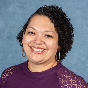 Portrait of female smiling. She has curly short hair with gold hoops and purple blouse. Blue Background.