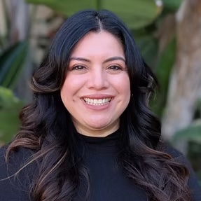 Selfie of female with a red top. She is smiling with Auburn hair. Her background is dark and not well lit. 