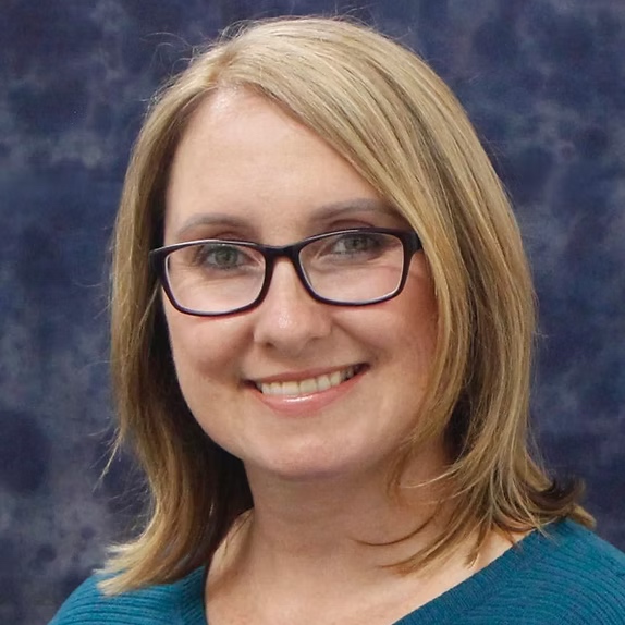 Photo of female blue top with blonde hair. She is smiling wearing black square glasses. The background is blue.