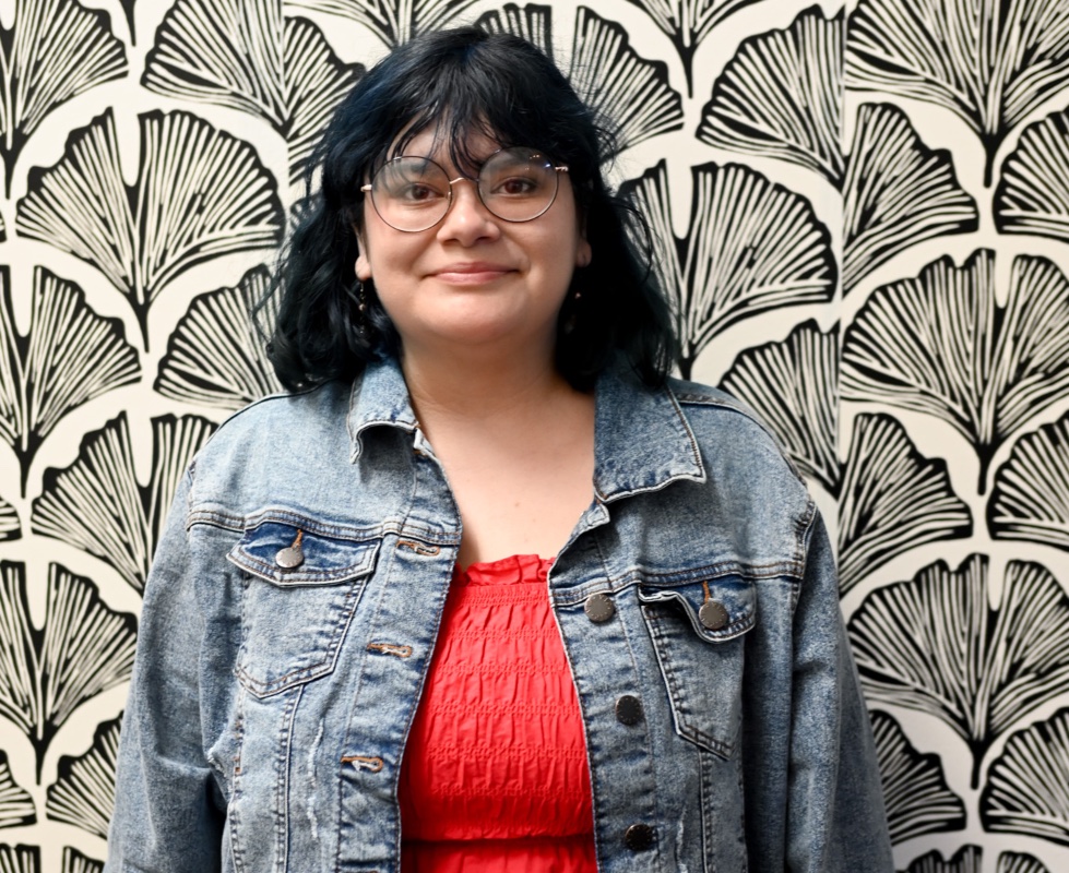 Female wearing jean jacket and red top behind a black and white floral design background. 