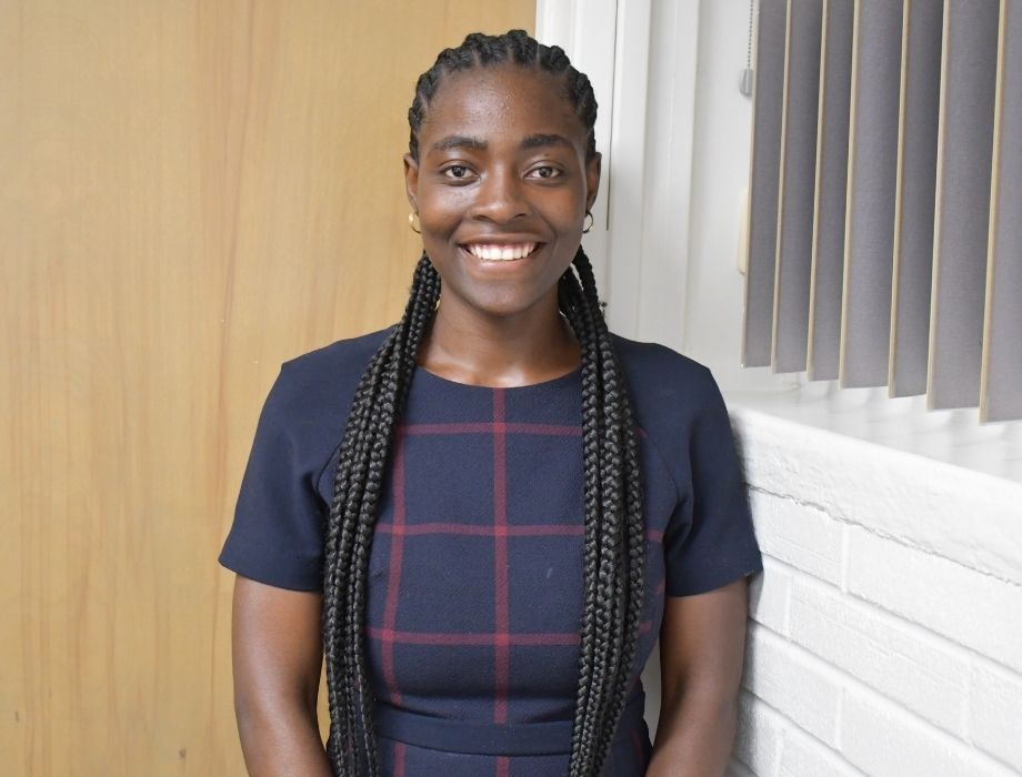 Female standing behind a white background. She has long braids and is smiling. 