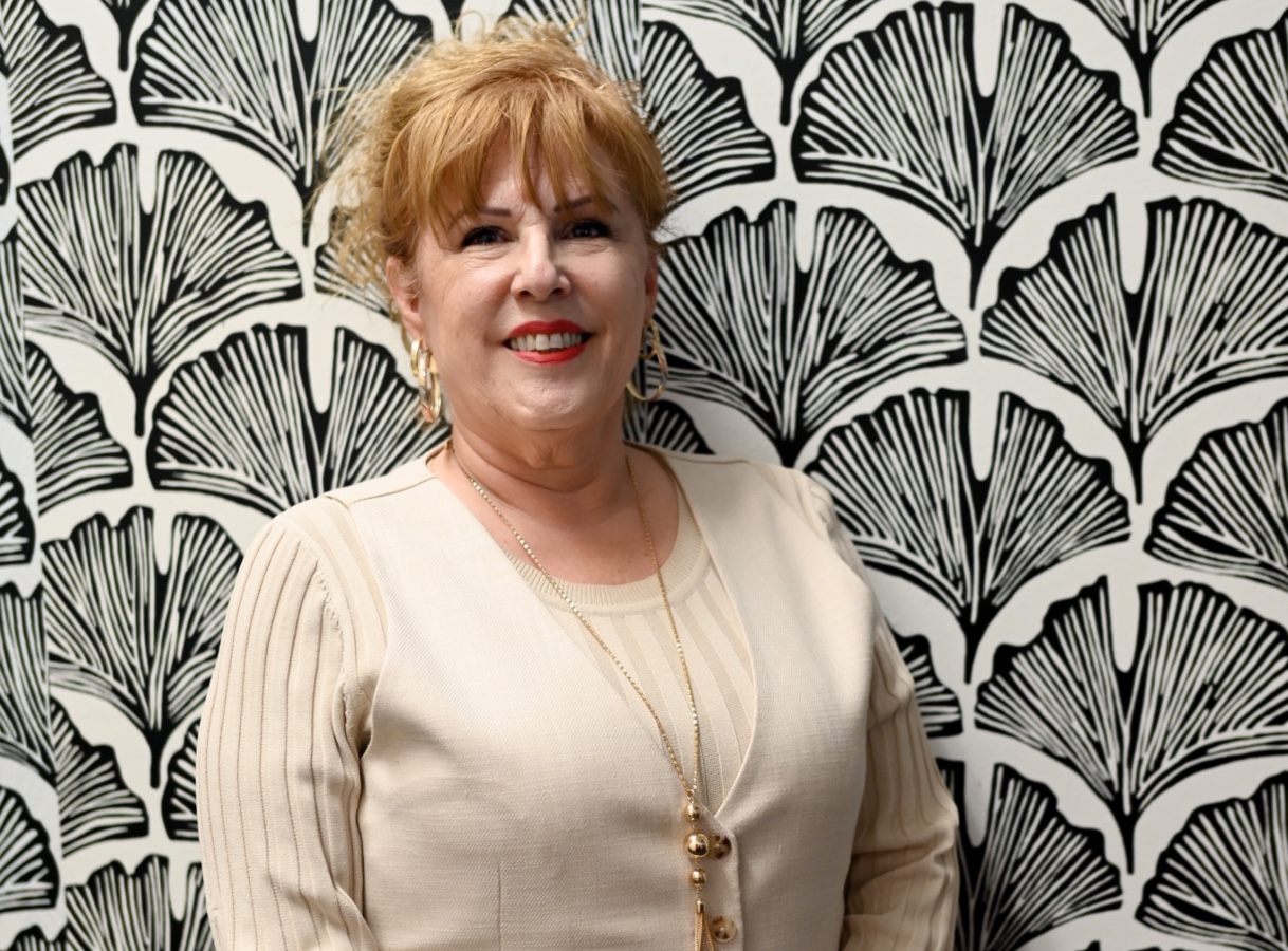 Female with blonde hair wearing a beige dress behind a black and white floral design background. 