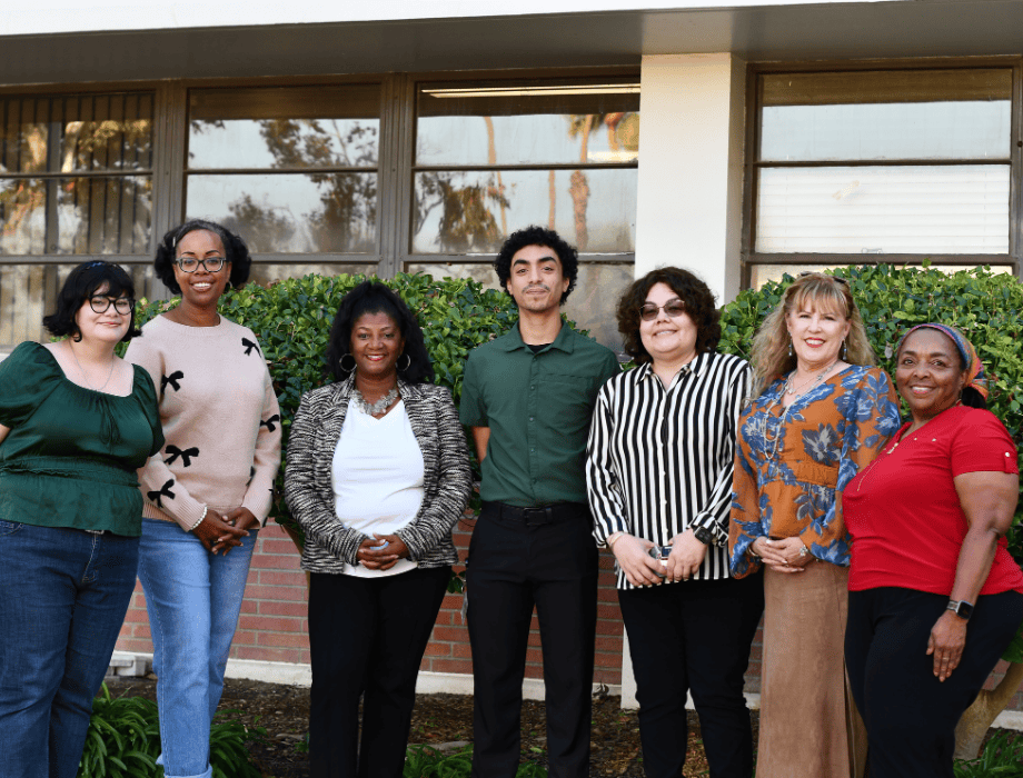 Group of students female and male standing and smiling in a row. There are two green bushes behind the. 