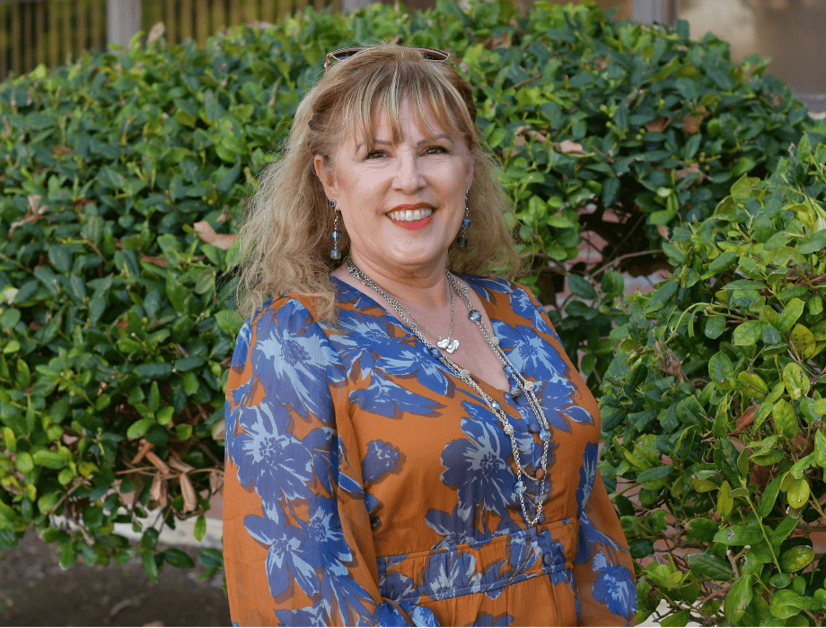 A female with blonde hair and is smiling. She has on a pattern shirt. Behind her are two green bushes.