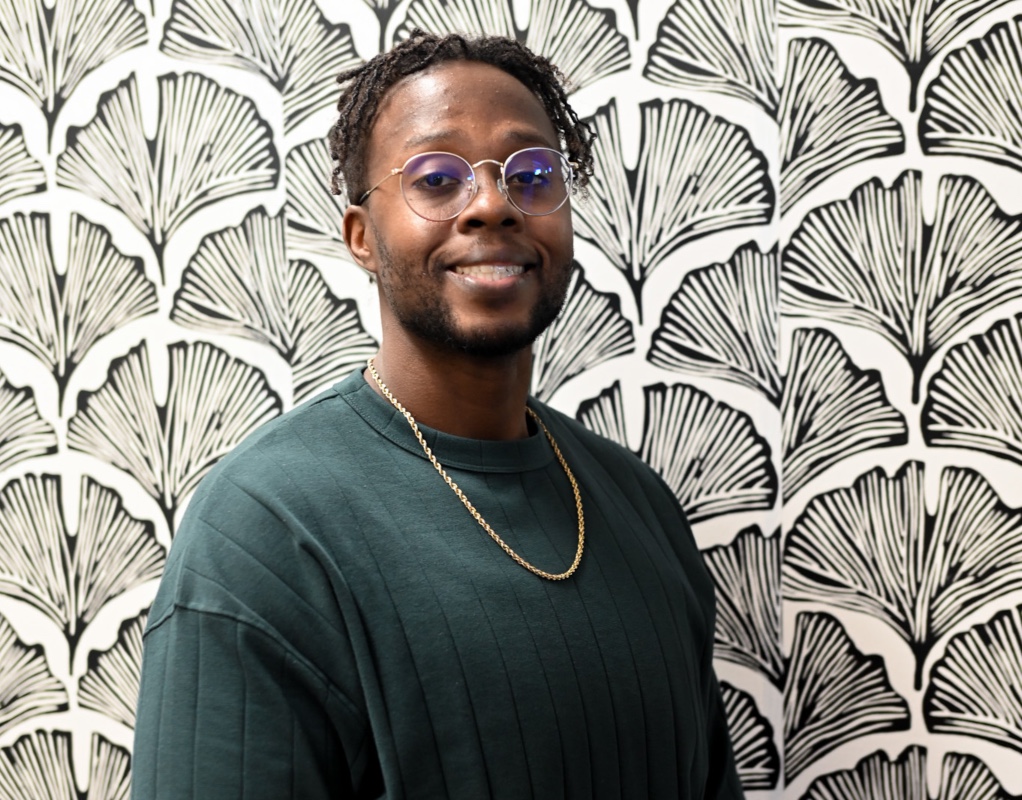 Male wearing green top with gold chain behind a black and white floral design background. 