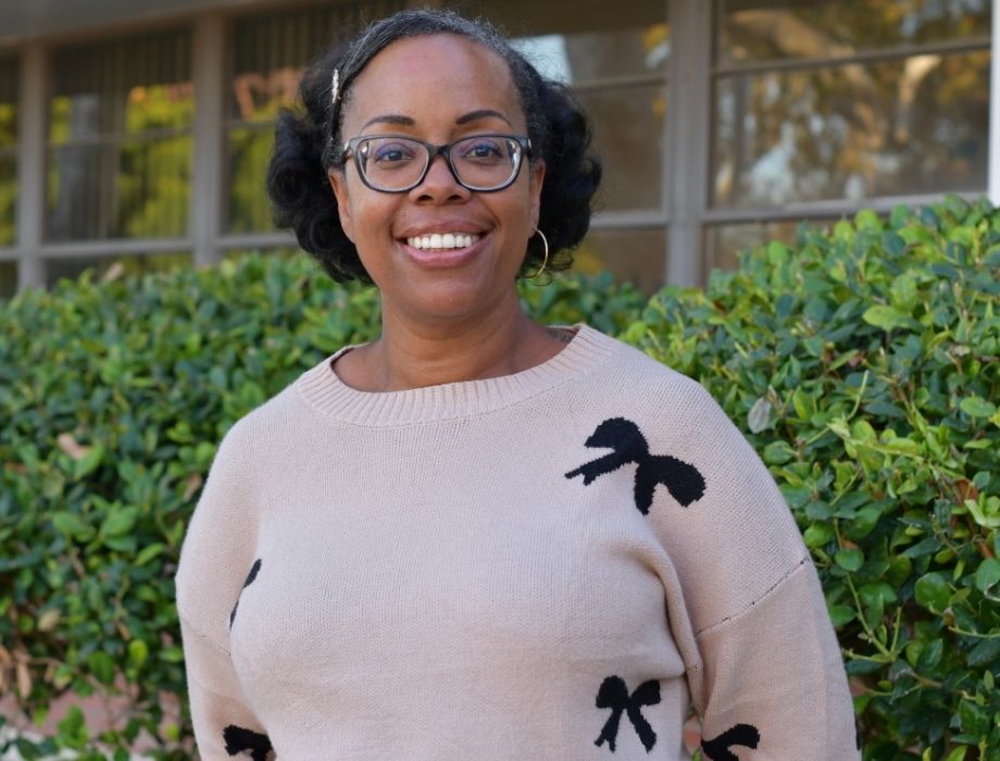 Female wearing black glasses her hair is pulled back. She is smiling wearing a tan sweater with black and white flowers. She is standing behind two green bushes. 