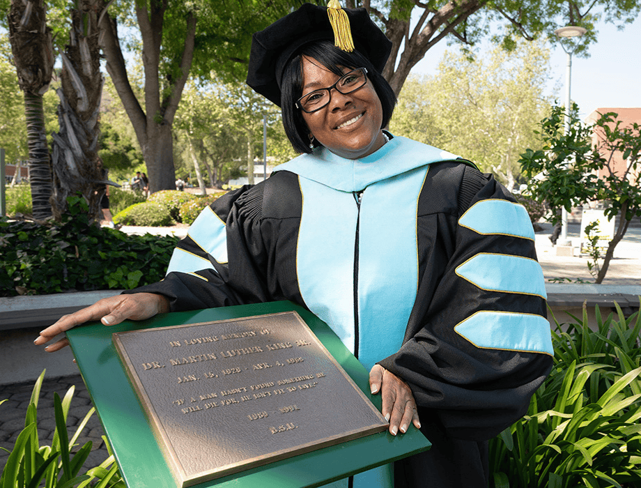 Female student in Regalia standing next to a plaque.