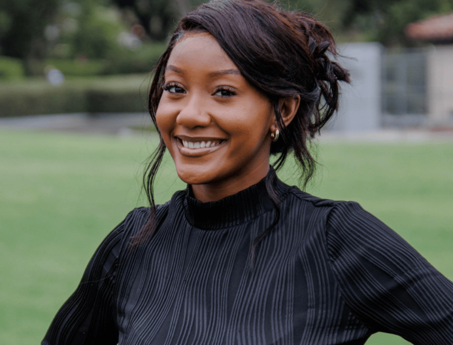 Female wearing a black turtleneck. She is smiling with her hair but up she is behind a grass lawn. 