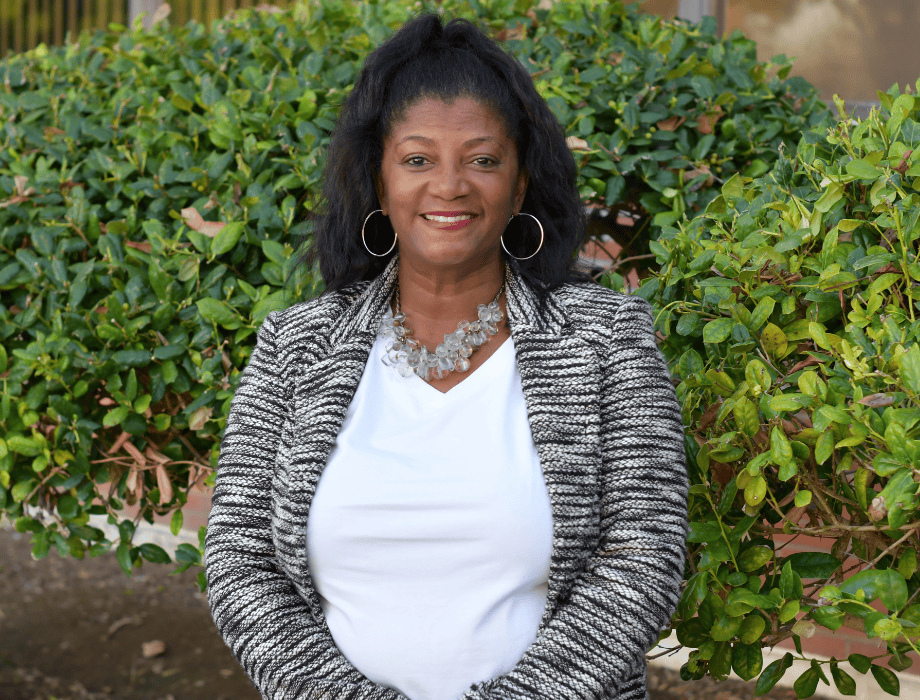 Female wearing a black and white pattern blazer and a white blouse. She has long black hair and is smiling. Behind her are two green bushes. 