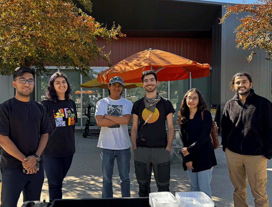 Students standing for a photo and smiling. They are behind a building and some trees during Fall. 