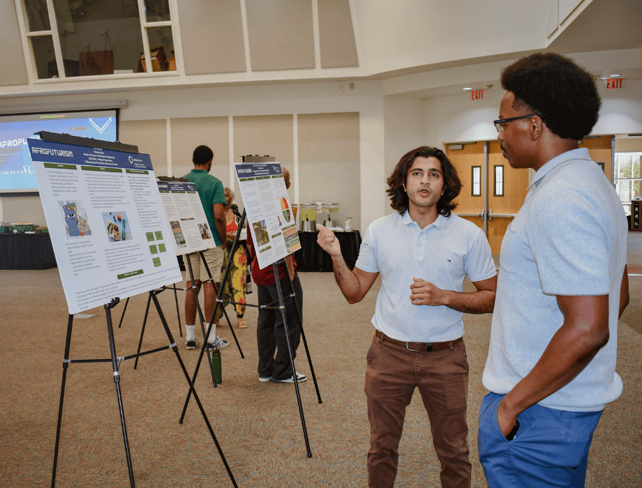 Male student explaining his project to another male student. There is a poster board next to them. 
