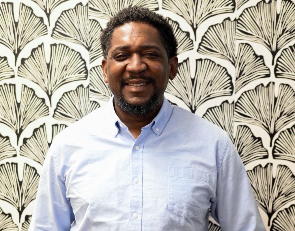 Male wearing blue top behind a black and white floral design background. 