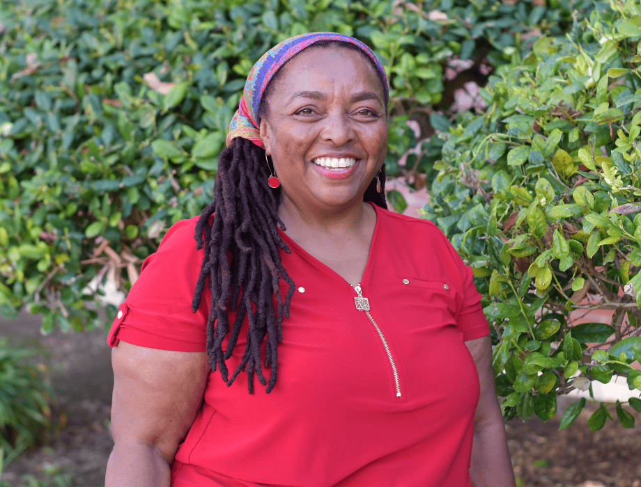 Female smiling with long brown hair and a red top. She is behind two green bushes.