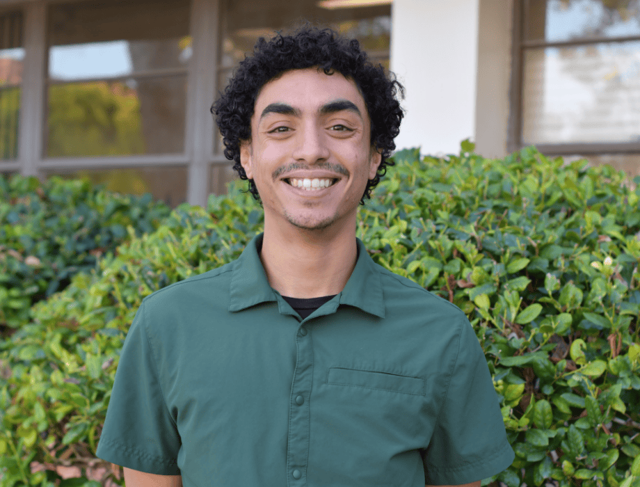 Male wearing a green button up with curly short hair and is smiling. He is behind two green bushes. 