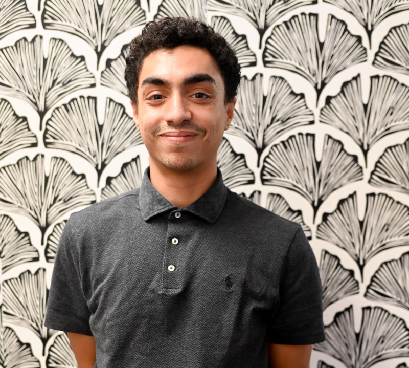 Male wearing grey behind a black and white floral design background. 