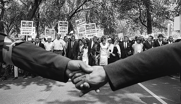 Marchers hold signs for jobs and equality; two people clasp hands in front.