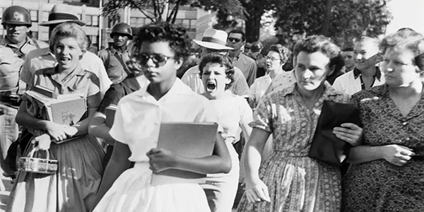 Young Black student walks with books, flanked by protesters and U.S. soldiers during 1957 desegregation.