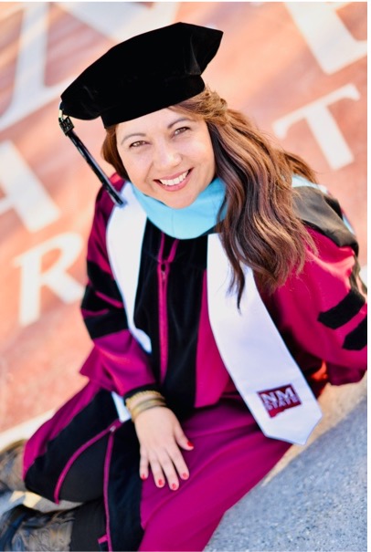 A woman with long brown hair wearing PhD robes and tam smiles at the camera
