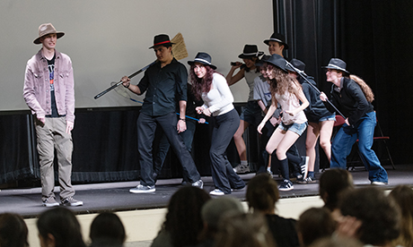 Performers onstage in hats sneaking behind a man, with an audience in front