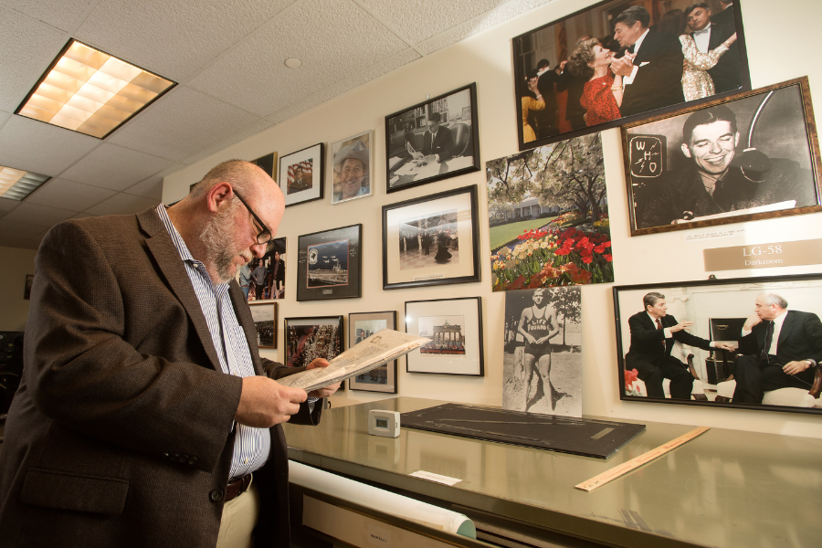 Ira Pemstein standing up examining a document in front of a collage of photos on a wall