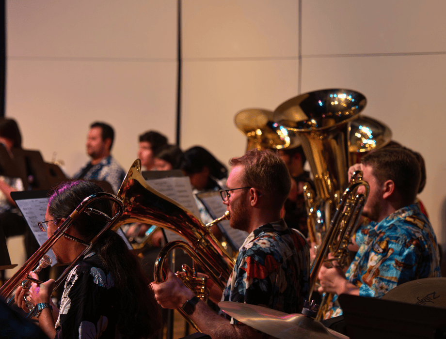 Brass musicians perform on stage, playing tubas and trombones from sheet music while seated in an ensemble setting.