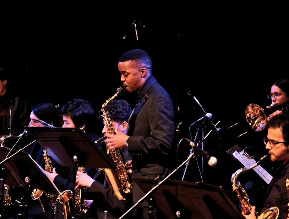 A saxophonist performs a solo on stage with a jazz ensemble seated behind music stands under stage lighting.