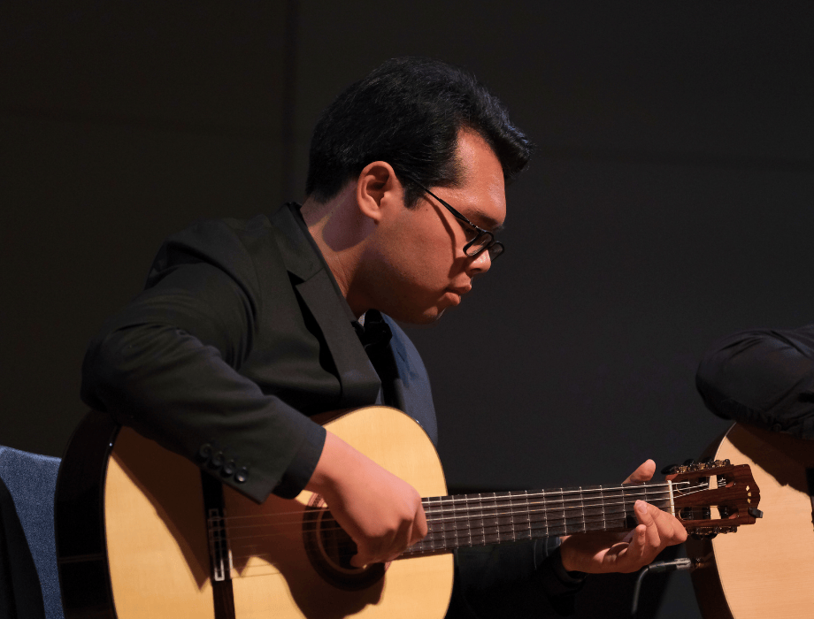 A guitarist in a dark suit plays a classical guitar on stage, focused on the fretboard under soft stage lighting.
