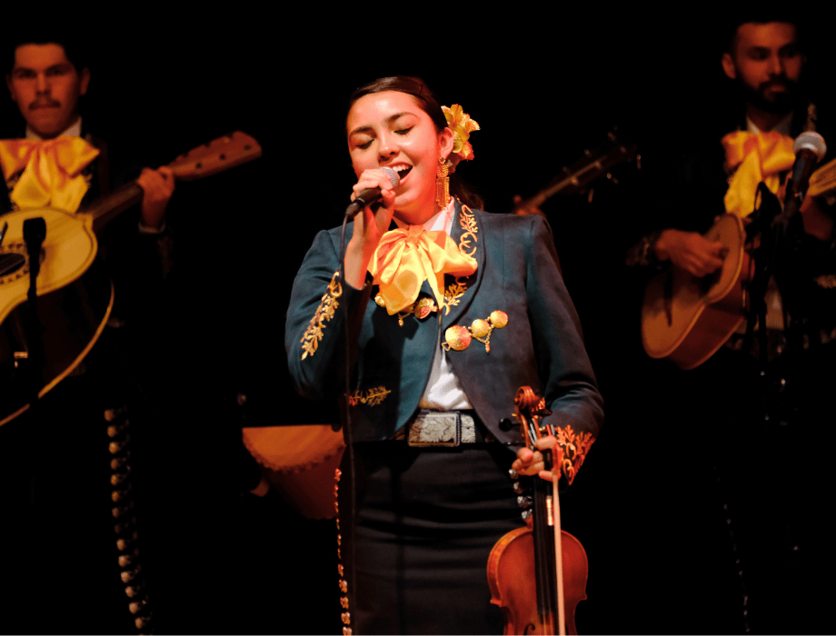 A mariachi singer performs on stage holding a violin, wearing a traditional charro outfit, with guitarists playing behind her.