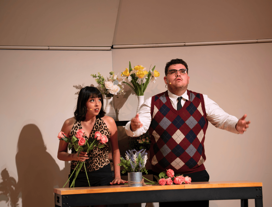 Two performers act a musical theater scene on stage, arranging flowers at a table while gesturing expressively.
