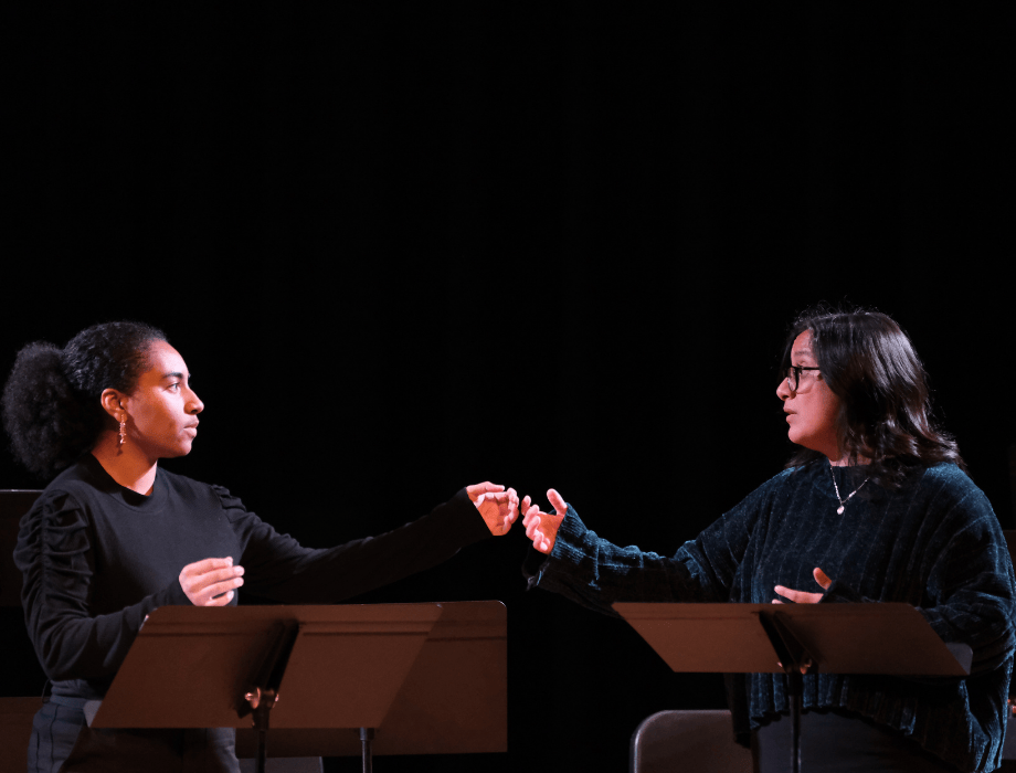 Two performers face each other on stage, gesturing expressively across music stands during a contemporary performance.