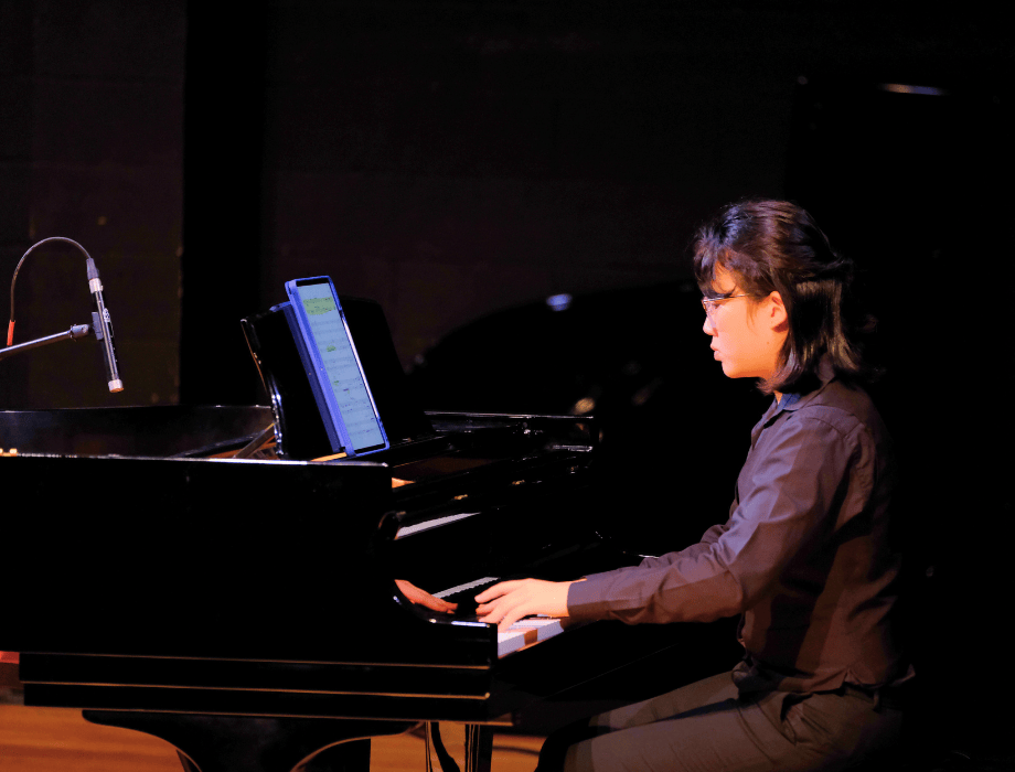 A pianist performs at a grand piano on stage, reading digital sheet music on a tablet under soft concert lighting.