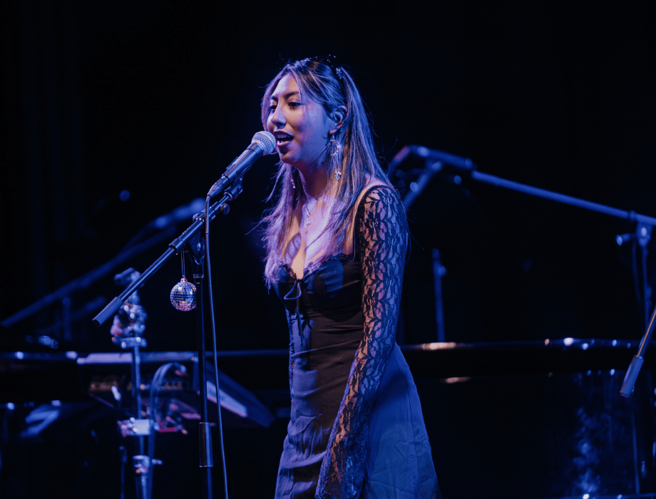 A singer performs into a microphone on a dimly lit stage, wearing a black dress with lace sleeves, with instruments and stands in the background.