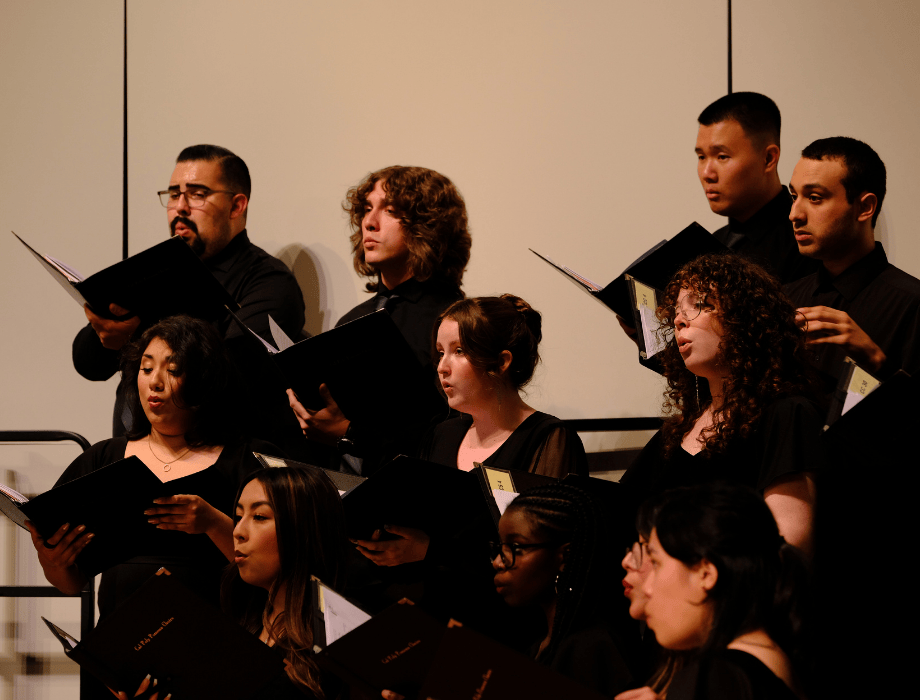 Members of the Cal Poly Pomona choirs perform on stage, singing from sheet music during a concert.