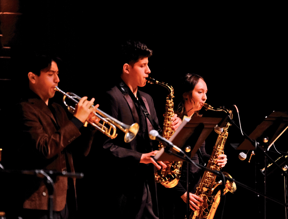 Student musicians perform in a jazz combo, playing trumpet and saxophones on stage during a concert.
