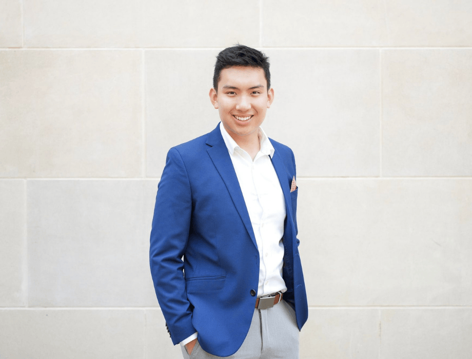 Portrait of composer Kevin Charoensri wearing a blue blazer and white shirt, smiling against a light stone wall background.