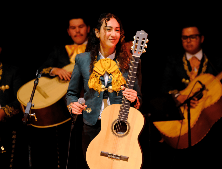 Members of Mariachi Los Broncos perform on stage, featuring a musician holding a guitar and smiling during the concert.