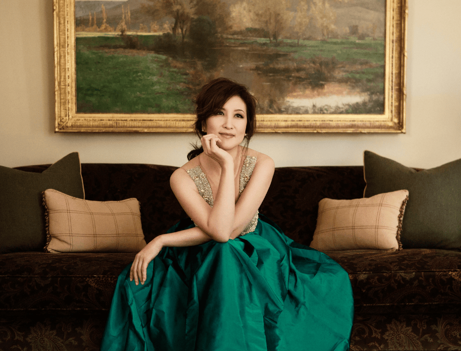 Pianist Min Kwon poses elegantly on a sofa wearing a green gown, with a framed landscape painting in the background.
