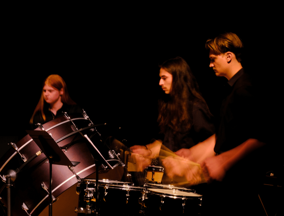 Student percussionists perform on stage, playing large drums and snare drums during a concert.